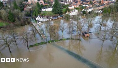 An aerial shot of a wide area of flooding on fields adjacent to a river. A bridge can be seen over the water and houses along the banks. Trees and a building are partially submerged.