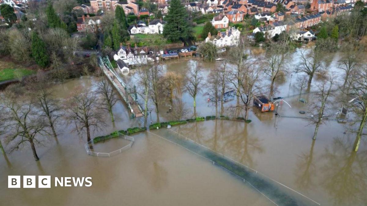 An aerial shot of a wide area of flooding on fields adjacent to a river. A bridge can be seen over the water and houses along the banks. Trees and a building are partially submerged.