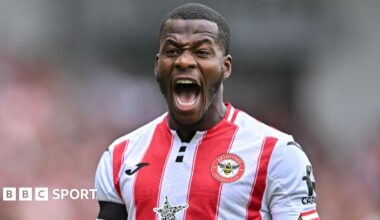 Dango Ouattara shown from chest up playing for Brentford Football Club in the home jersey with wide red and white vertical stripes. The forward is opening his mouth very wide to shout, with his teeth and tongue visible