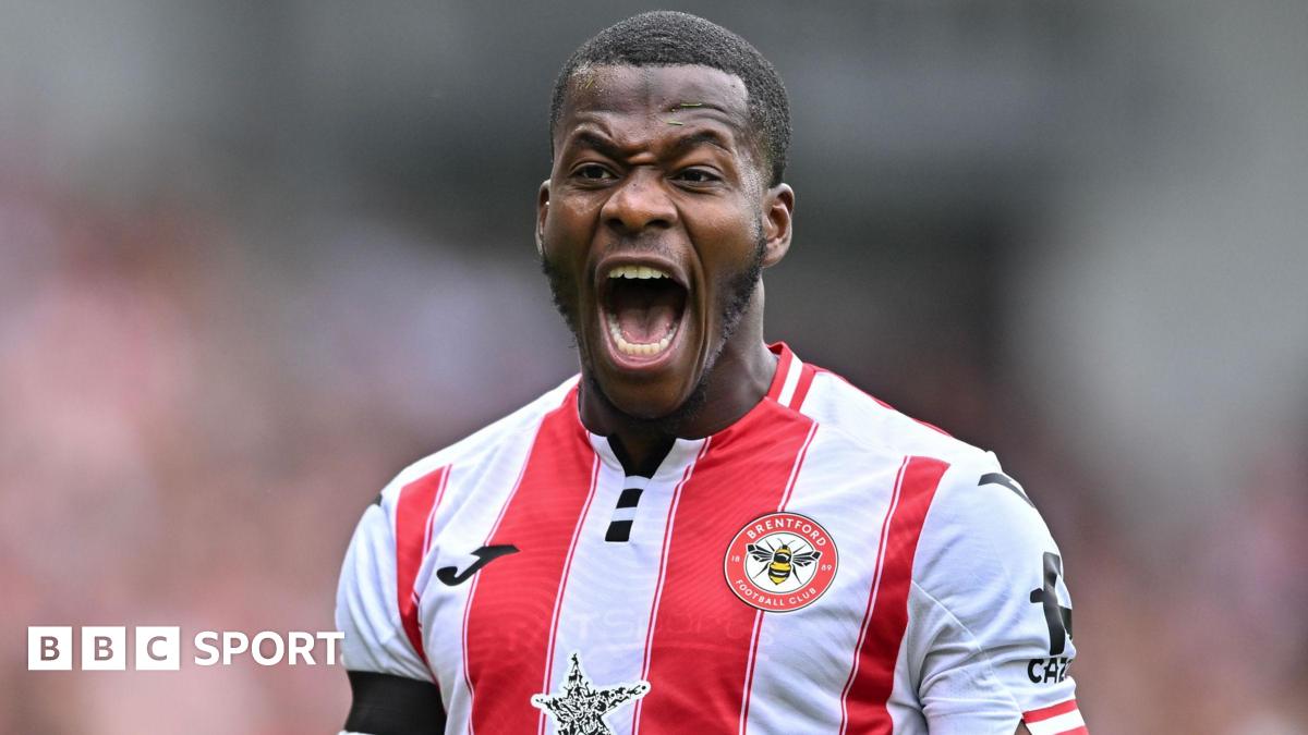 Dango Ouattara shown from chest up playing for Brentford Football Club in the home jersey with wide red and white vertical stripes. The forward is opening his mouth very wide to shout, with his teeth and tongue visible