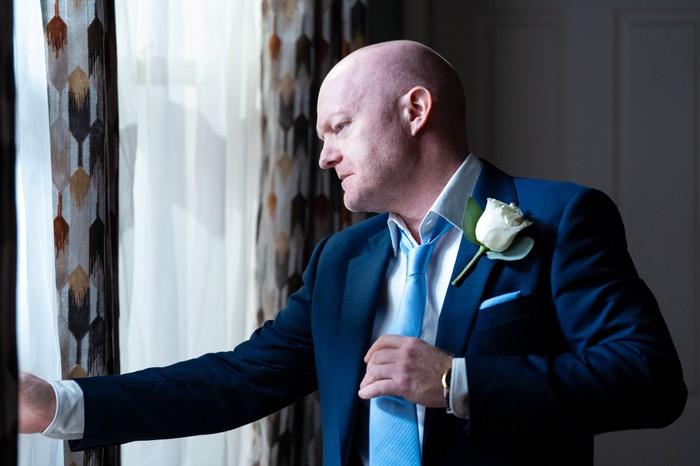 Max wears a navy suit and a white rose buttonhole as he stares out of the window in EastEnders