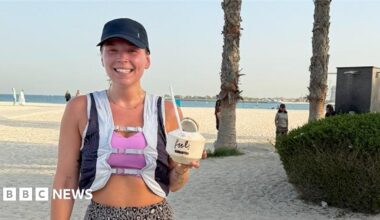 Sol Hyde takes a selfie. He is sitting on a bench with his laptop. He is wearing a black T-shirt, khaki trousers and white trainers. He has white earphones in. There are autumn leaves on the ground and a number of Lime hire bikes behind him.