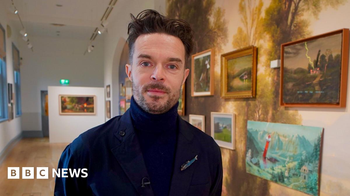 Oliver Jeffers is a man with black hair and beard who is standing in an art gallery in front of a wall of artworks. He is wearing a navy turtleneck top and coat.