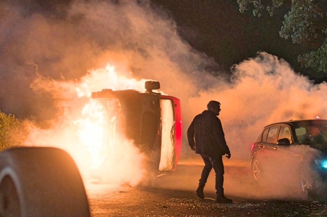 An undisclosed person surrounded by crashed, flaming and smoking cars at night, in a still from Corriedale.