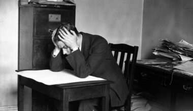 Pessimistic businessman sitting at a desk with a frown, surrounded by office documents and a computer, conveying workplace...