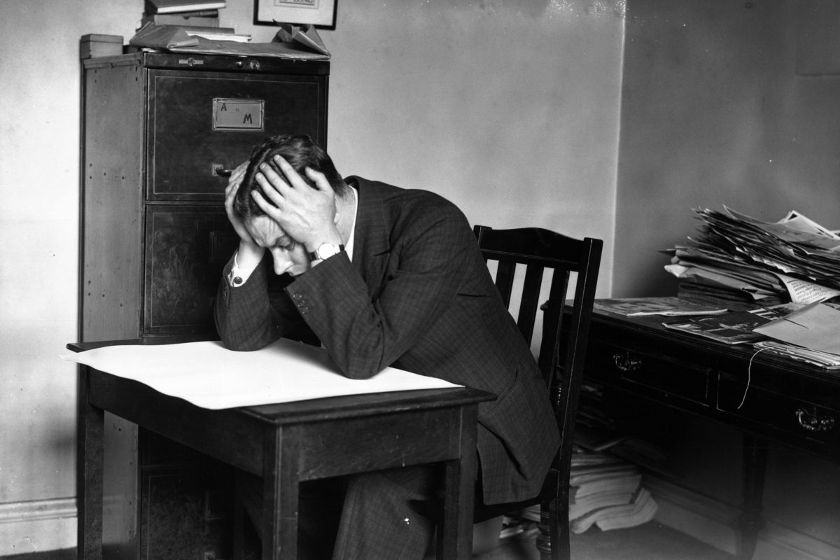 Pessimistic businessman sitting at a desk with a frown, surrounded by office documents and a computer, conveying workplace...