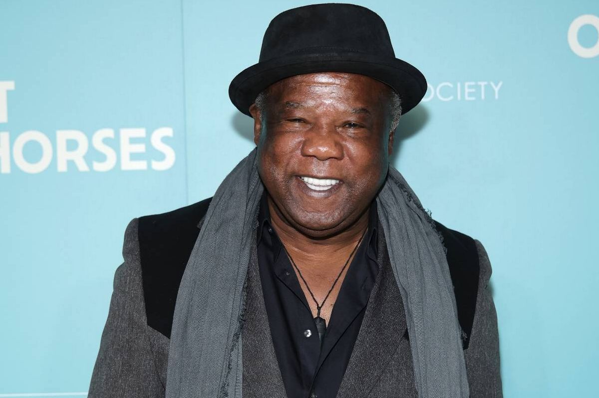 An individual dressed in a black hat, a gray scarf, and a dark jacket poses for a photograph against a blue backdrop with the word "Horses" prominently displayed.