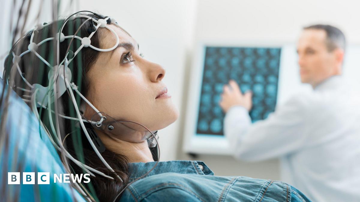 Woman lying on a medical bed wearing a cap with multiple electrodes and wires attached to the head. In the background, a healthcare professional in a white coat is examining brain scan images displayed on a monitor.