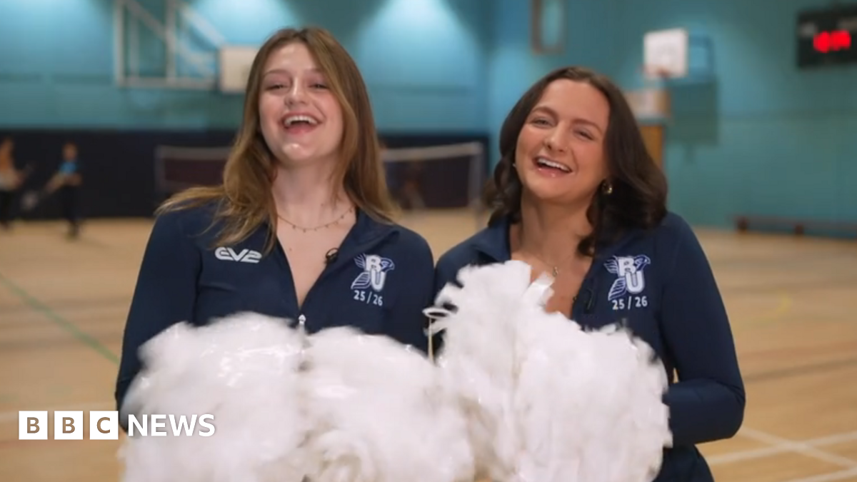 Eve Kimberley (left) and Liv Teague are wearing dark blue cheerleaders outfits and waving pompoms in a gym.