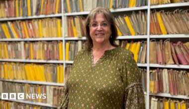 A woman with dark shoulder length hair and glasses smiles for the camera. She is wearing a dark green flowered top in front of a white shelving unit with colourful paper files