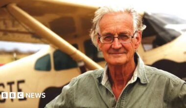 A brown NC500 sign post which says it is 27 miles to Ullapool next to a country road with hills in the background.