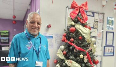An older gentleman, in his 70s, stands smiling in front of a beautifully adorned Christmas tree and a notice board pinned with cards. He is South Asian. He wears a blue t shirt and a lanyard, both of which identify him as an NHS volunteer. The space around him is festooned with Christmas decorations.