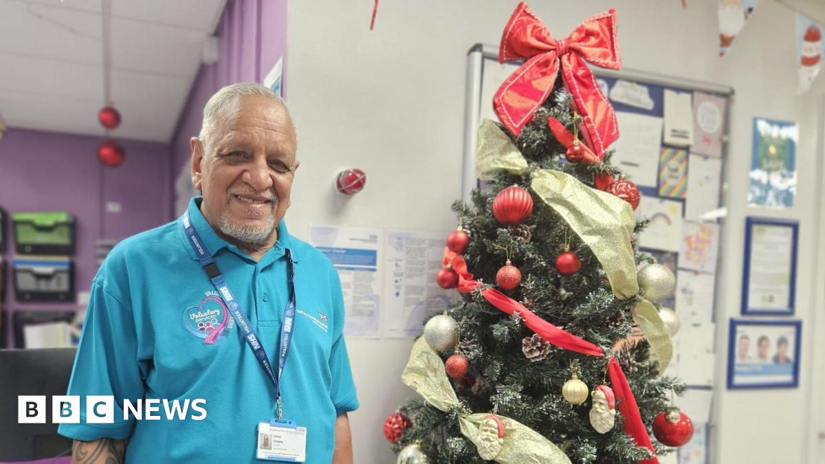 An older gentleman, in his 70s, stands smiling in front of a beautifully adorned Christmas tree and a notice board pinned with cards. He is South Asian. He wears a blue t shirt and a lanyard, both of which identify him as an NHS volunteer. The space around him is festooned with Christmas decorations.