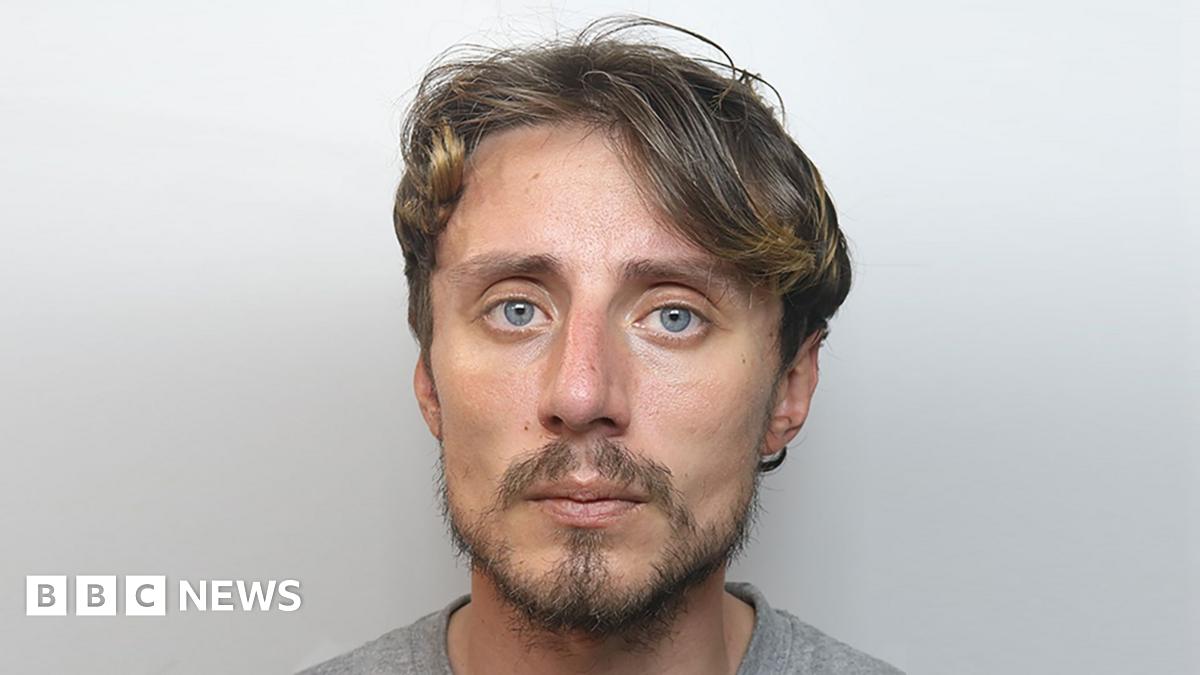 A mugshot photo of a man with a light darkly-coloured beard and dark hair