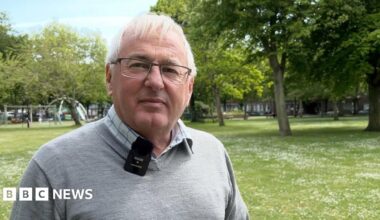 Kevin Keen has grey hair and glasses and is smiling at the camera. He is wearing a grey jumper and a chequered blue and white shirt.