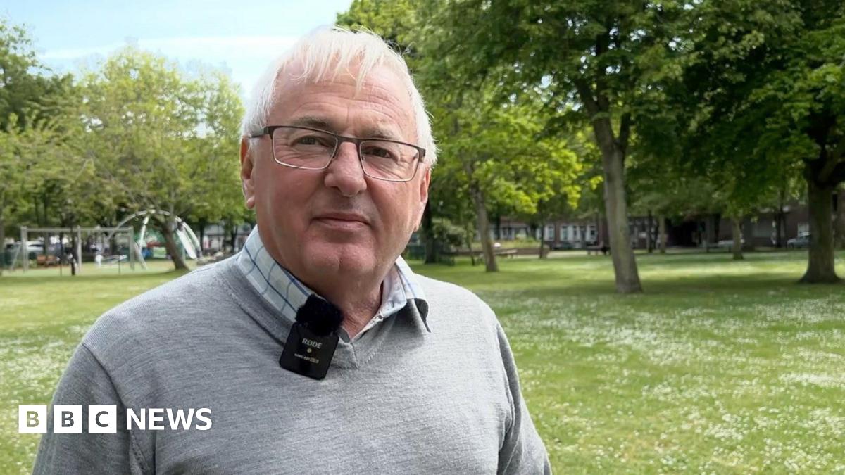 Kevin Keen has grey hair and glasses and is smiling at the camera. He is wearing a grey jumper and a chequered blue and white shirt.