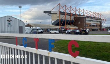 A white gate with the letters I C T F C in red and blue writing in front of a brick football stand with orange pillars. Various cars are in the foreground of a car park, as well as a grassy area to the right of the picture.