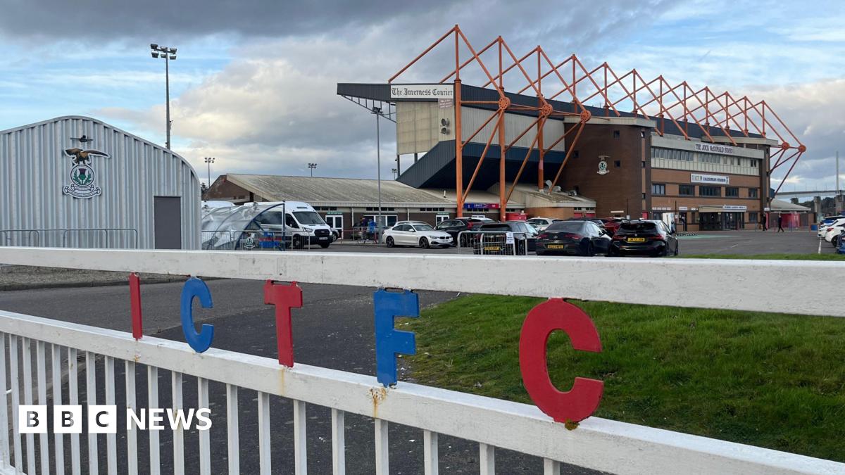 A white gate with the letters I C T F C in red and blue writing in front of a brick football stand with orange pillars. Various cars are in the foreground of a car park, as well as a grassy area to the right of the picture.