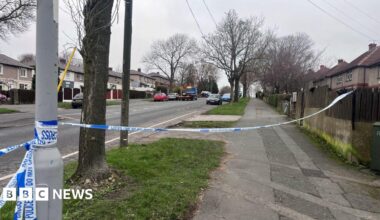 Police tape reading “POLICE LINE DO NOT CROSS” stretches across a suburban street, blocking access. Houses, parked cars, and bare trees line the road in the background on an overcast day.