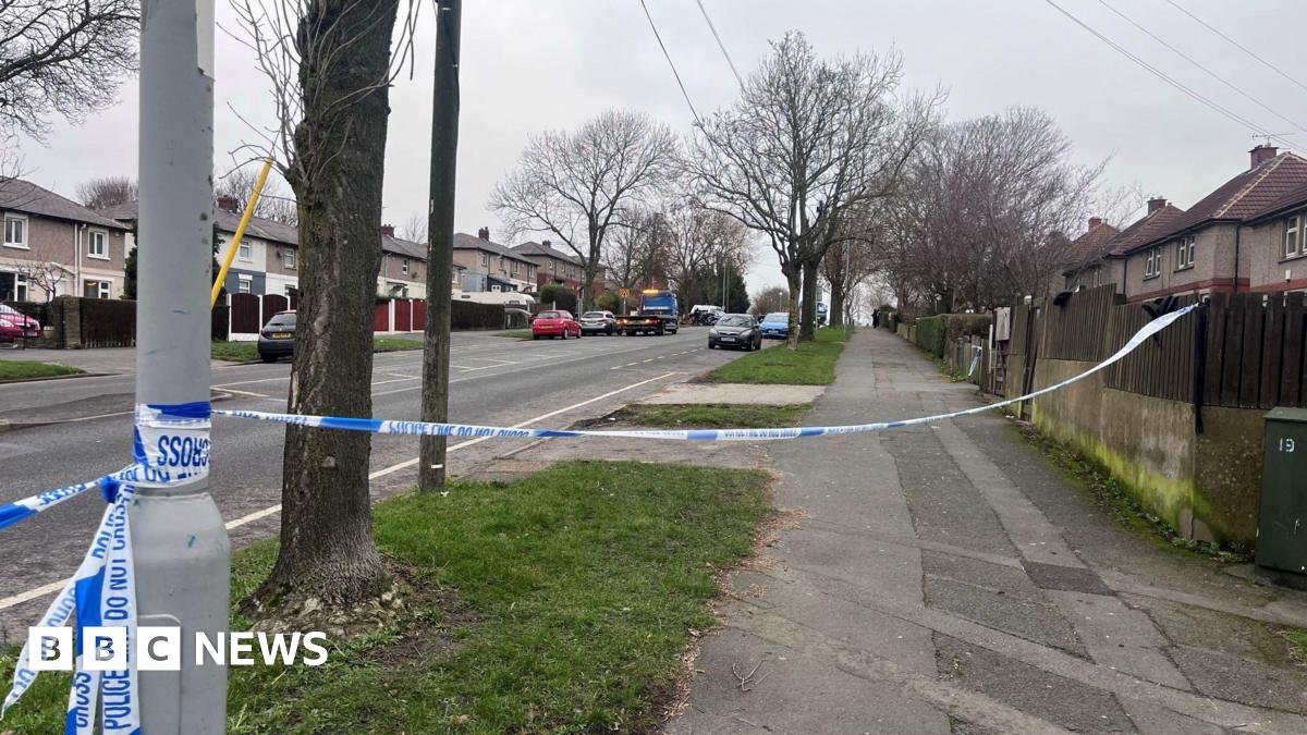 Police tape reading “POLICE LINE DO NOT CROSS” stretches across a suburban street, blocking access. Houses, parked cars, and bare trees line the road in the background on an overcast day.