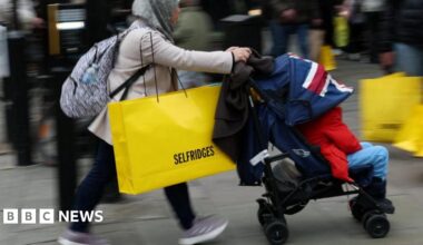 A woman wearing a hijab pushes a pram while carrying a large yellow Selfridges bag down the street.