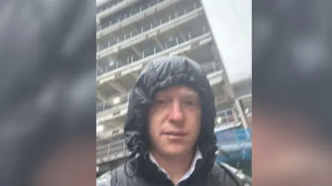 Sol Hyde Sol with a hood up in the rain in front of a high-rise building.