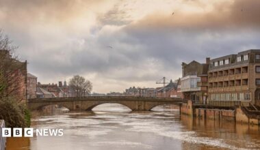 A 19th century stone bridge spanning a river swollen with flood water. There are buildings on each side and a cloudy sky above.