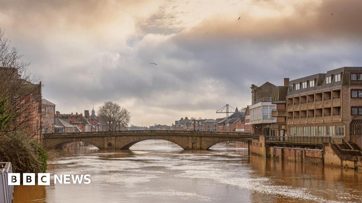 A 19th century stone bridge spanning a river swollen with flood water. There are buildings on each side and a cloudy sky above.