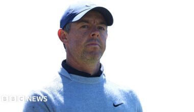 Rory McIlroy, a man with short, greying dark hair, pictured outdoors with green trees in the background.  He wears a black cap with a white tick in the centre. He wears a black polo top and holds a white golf glove in his left hand.