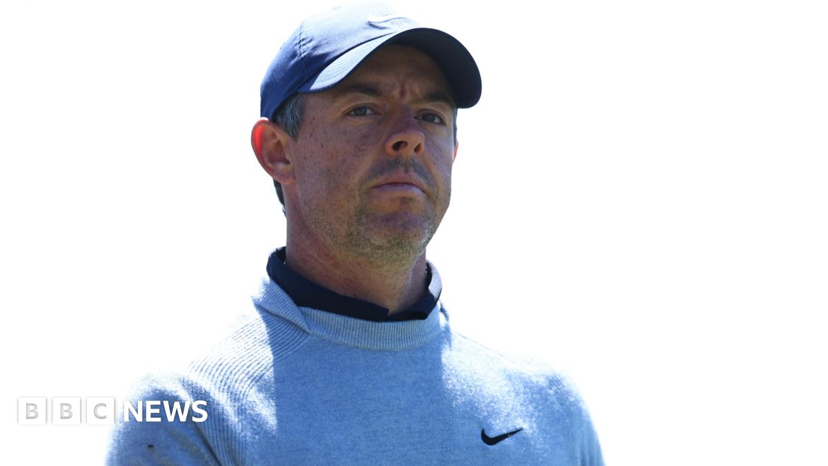 Rory McIlroy, a man with short, greying dark hair, pictured outdoors with green trees in the background.  He wears a black cap with a white tick in the centre. He wears a black polo top and holds a white golf glove in his left hand.