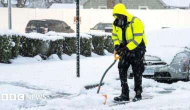A man in a fluorescent jacket and black trousers and boots shovels snow by the side of a road
