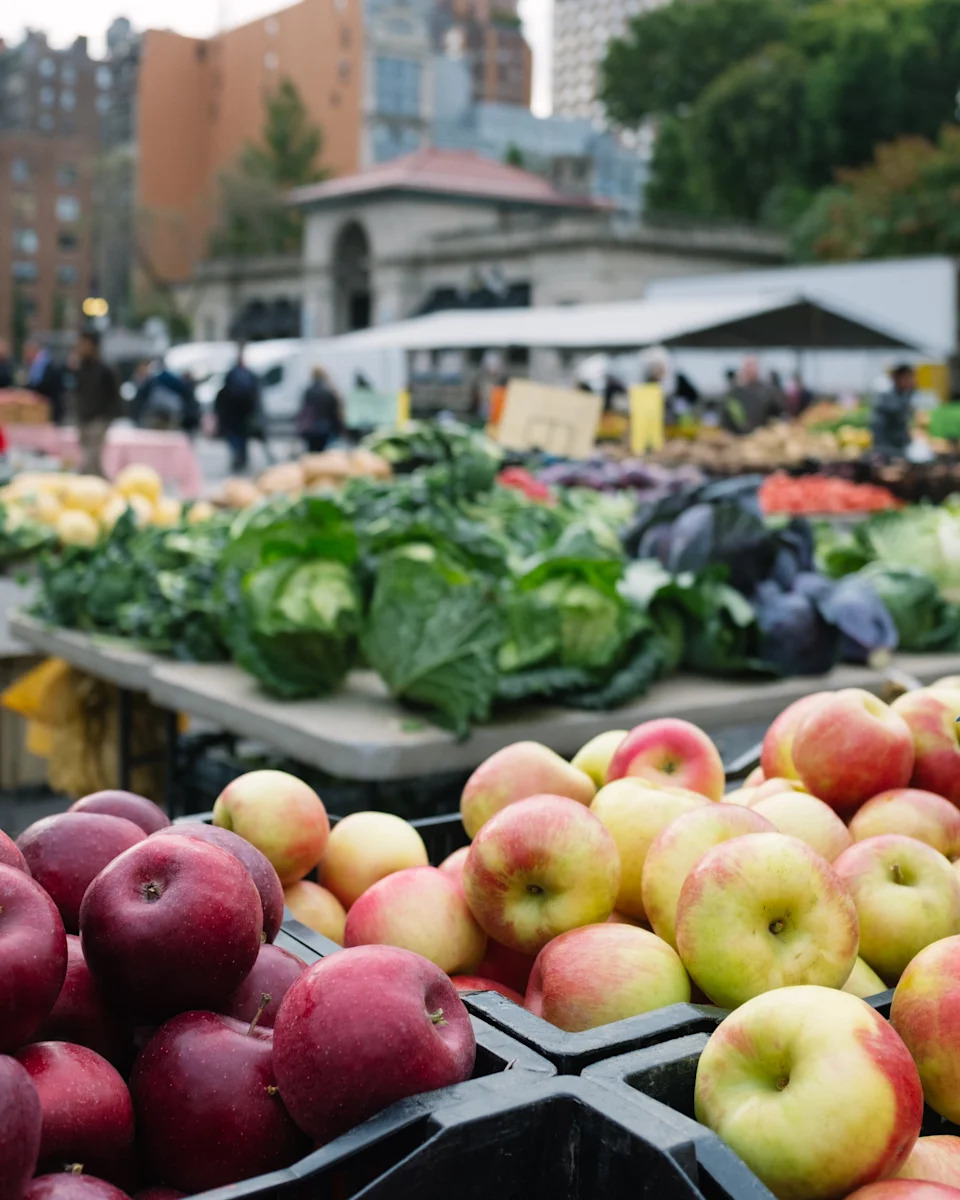 Fresh produce, including apples and leafy greens, is displayed at an outdoor market in an urban setting