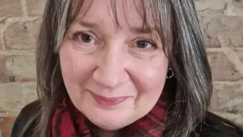 Deborah Heneghan A head and shoulders image of Deborah Heneghan, a woman with brown, shoulder-length hair streaked with grey. She is smiling at the camera, wearing a red and black checked scarf and silver earrings.