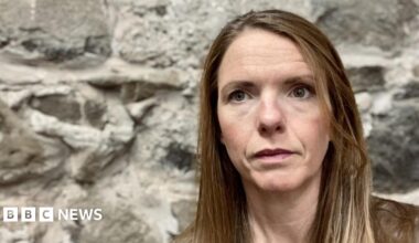 A headshot of Victoria Nichols, who has long brown hair, she is sitting in front of a grey stone wall and is looking slightly off to the left of the camera.