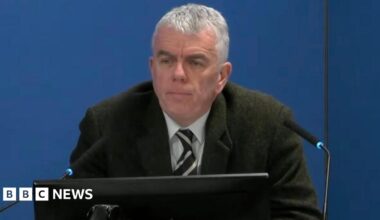 A man with tie and dark jacket sits in front of two microphones and a monitor