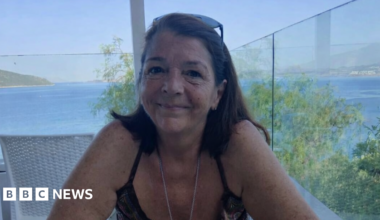 Julie Wilson is pictured with shoulder-length brown hair sitting at a table on a terrace overlooking the sea, smiling at the camera, with blue water and a distant coastline visible behind her.