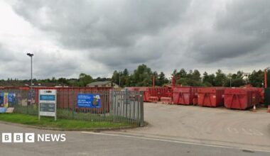 The entrance to a household recycling centre on a grey day. large metal fencing around it with huge red metal containers for recycling.
