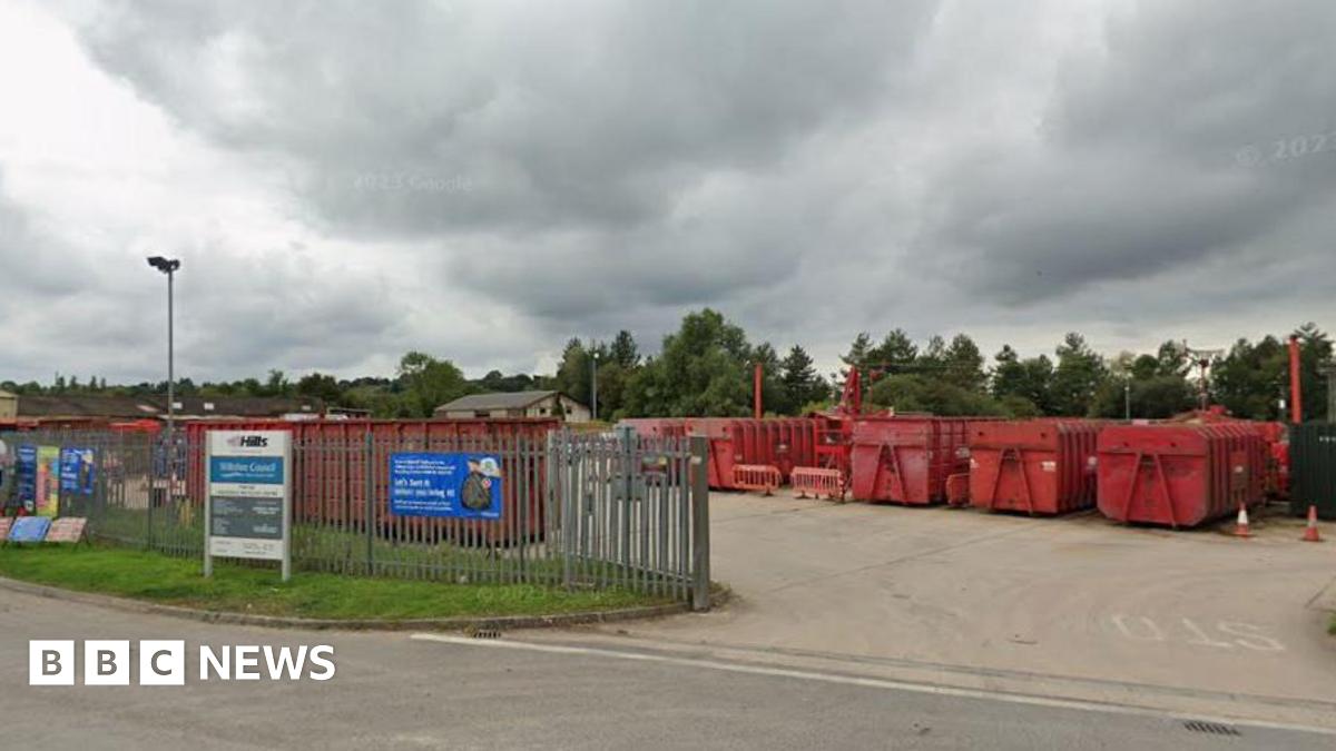 The entrance to a household recycling centre on a grey day. large metal fencing around it with huge red metal containers for recycling.