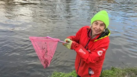 Caroline Brice pictured next to the River Irwell holding a fishing net. She is wearing a red coat.