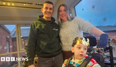 Dave and Emma stand with their arms around each other behind their son who is in a wheelchair. He has a gold grown on and it wearing a bright stripy jumper. Dave is wearing a green hoody and has dark brown hair and Emma is wearing a grey sparkly jumper and has blonde hair.