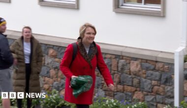 Deputy Sasha Kazantseva-Miller - A woman with blonde hair walking up a hill, wearing a red coat and a back pack. In her right hand she is carrying a green carrier bag.