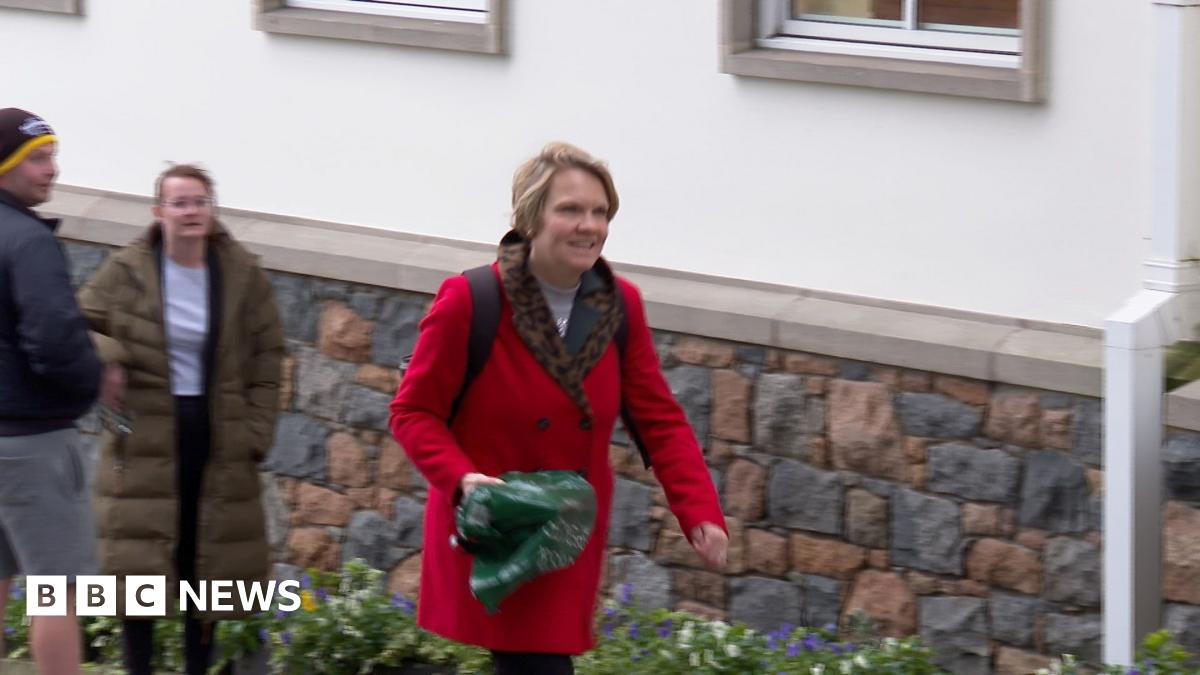 Deputy Sasha Kazantseva-Miller - A woman with blonde hair walking up a hill, wearing a red coat and a back pack. In her right hand she is carrying a green carrier bag.