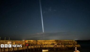 A cobblestone harbour with a small white paddle boat docked, and a dark blue morning sky with stars and a bright white vertical light shining from the sea upwards.
