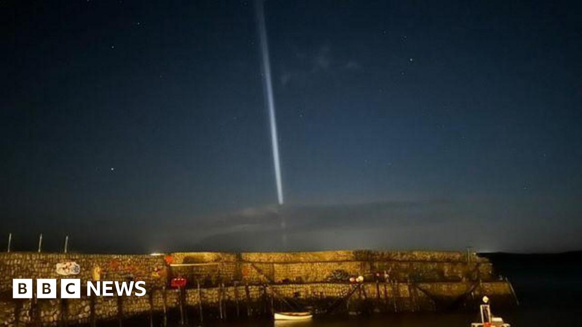 A cobblestone harbour with a small white paddle boat docked, and a dark blue morning sky with stars and a bright white vertical light shining from the sea upwards.