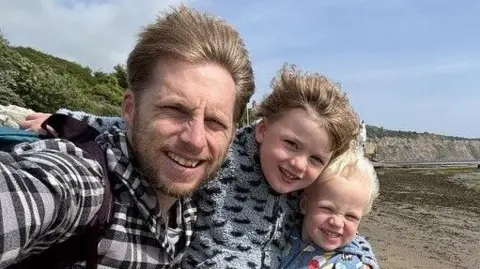 Andrew Andrew with his two young sons stand on a beach and pose for a selfie. Andrew has light brown hair and a beard and is wearing a checked shirt. The boys are both wearing fleeces. Their hair is being swept back by the wind