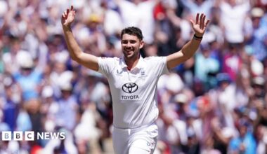 Josh Tongue wearing a white England cricket kit with the crest over his left breast and the Toyota logo on his stomach. He raises his arms in the air with a crowd in the background.