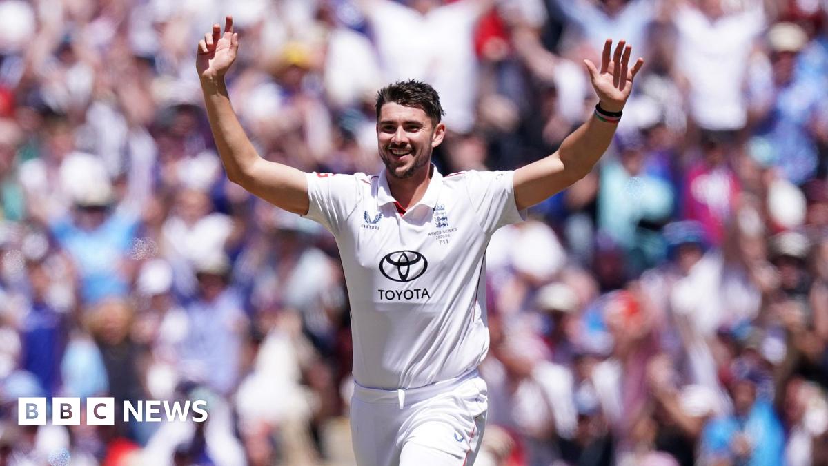 Josh Tongue wearing a white England cricket kit with the crest over his left breast and the Toyota logo on his stomach. He raises his arms in the air with a crowd in the background.