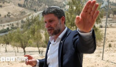 Israeli Finance Minister Bezalel Smotrich wears a white dress shirt and black suit jacket. He has short salt-and-pepper hair and beard. He is speaking and gesturing with his hands. Dry ground, shrubbery and a road winding through hills in the West Bank are seen behind him.