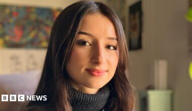 Aisha Belarbi sitting at a desk and turning to look directly down the camera. She is wearing a black leather jacket with a dark grey turtle neck sweater underneath. In her left hand she is holding a tablet computer. She is smiling, though her teeth are not showing. Her straight dark brown hair is parted in the middle of her head and is worn down, falling below her shoulders. On the right of the image is a large window, through which bright sunlight is entering the room. There is a lamp on the desk and the large green leaves of a houseplant can be seen growing up from the desk and towards the window.