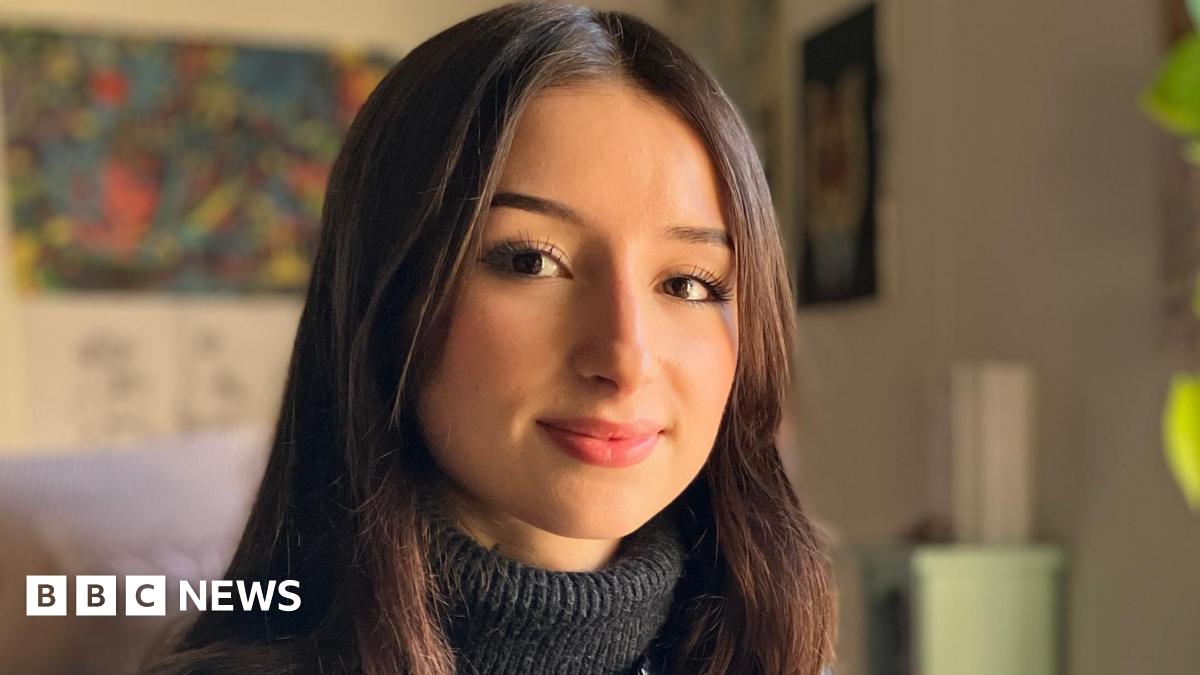 Aisha Belarbi sitting at a desk and turning to look directly down the camera. She is wearing a black leather jacket with a dark grey turtle neck sweater underneath. In her left hand she is holding a tablet computer. She is smiling, though her teeth are not showing. Her straight dark brown hair is parted in the middle of her head and is worn down, falling below her shoulders. On the right of the image is a large window, through which bright sunlight is entering the room. There is a lamp on the desk and the large green leaves of a houseplant can be seen growing up from the desk and towards the window.